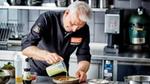 Een mannelijke chef staat in de keuken en giet saus over een gerecht.