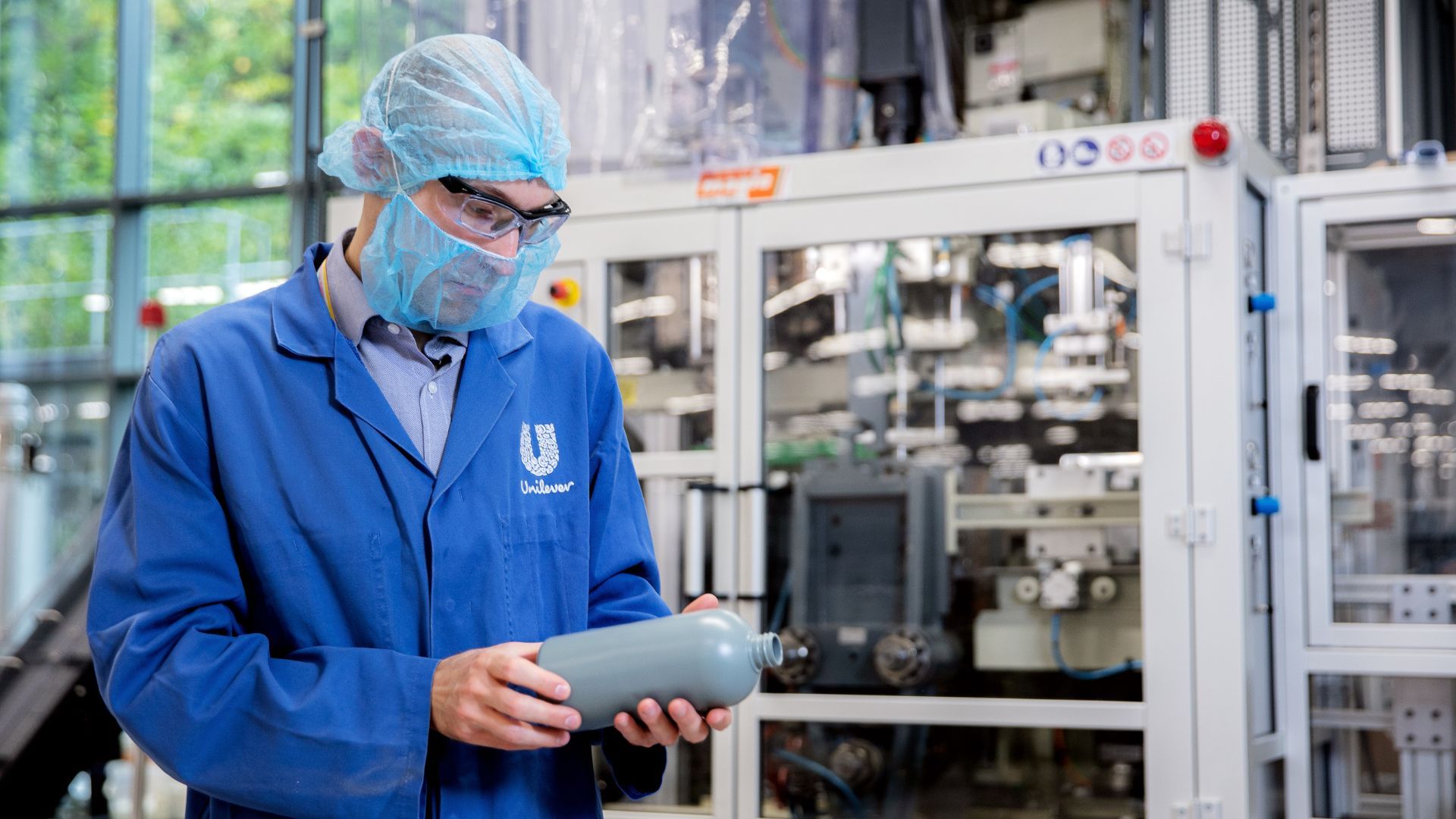 Een persoon in een blauwe Unilever-labjas, haarnetje, veiligheidsbril en mondkapje onderzoekt een grijze plastic fles in een fabrieksomgeving, met industriële apparatuur op de achtergrond.