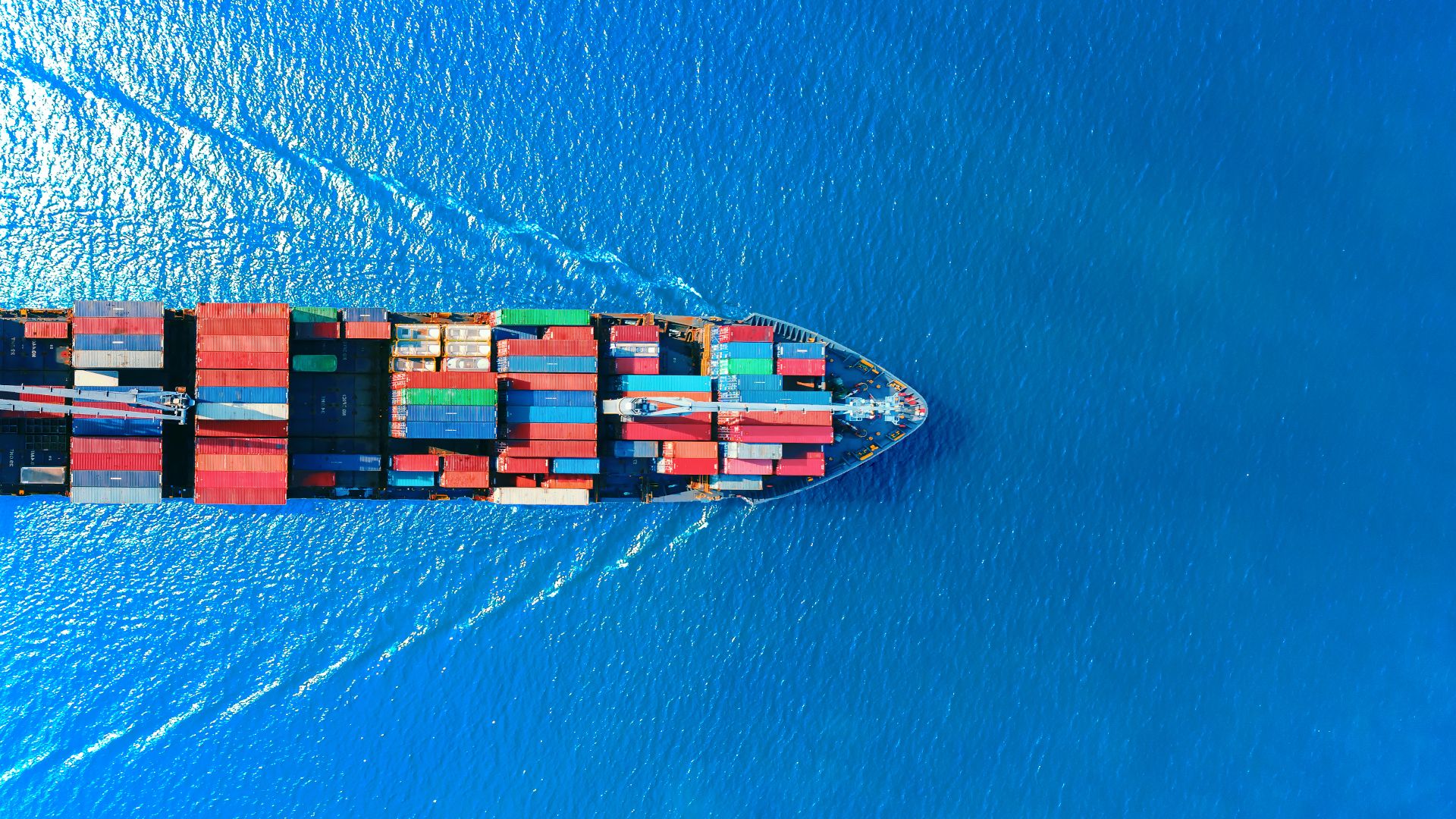 Een afbeelding van een boot van bovenaf, geladen met zeecontainers, varend op open water.