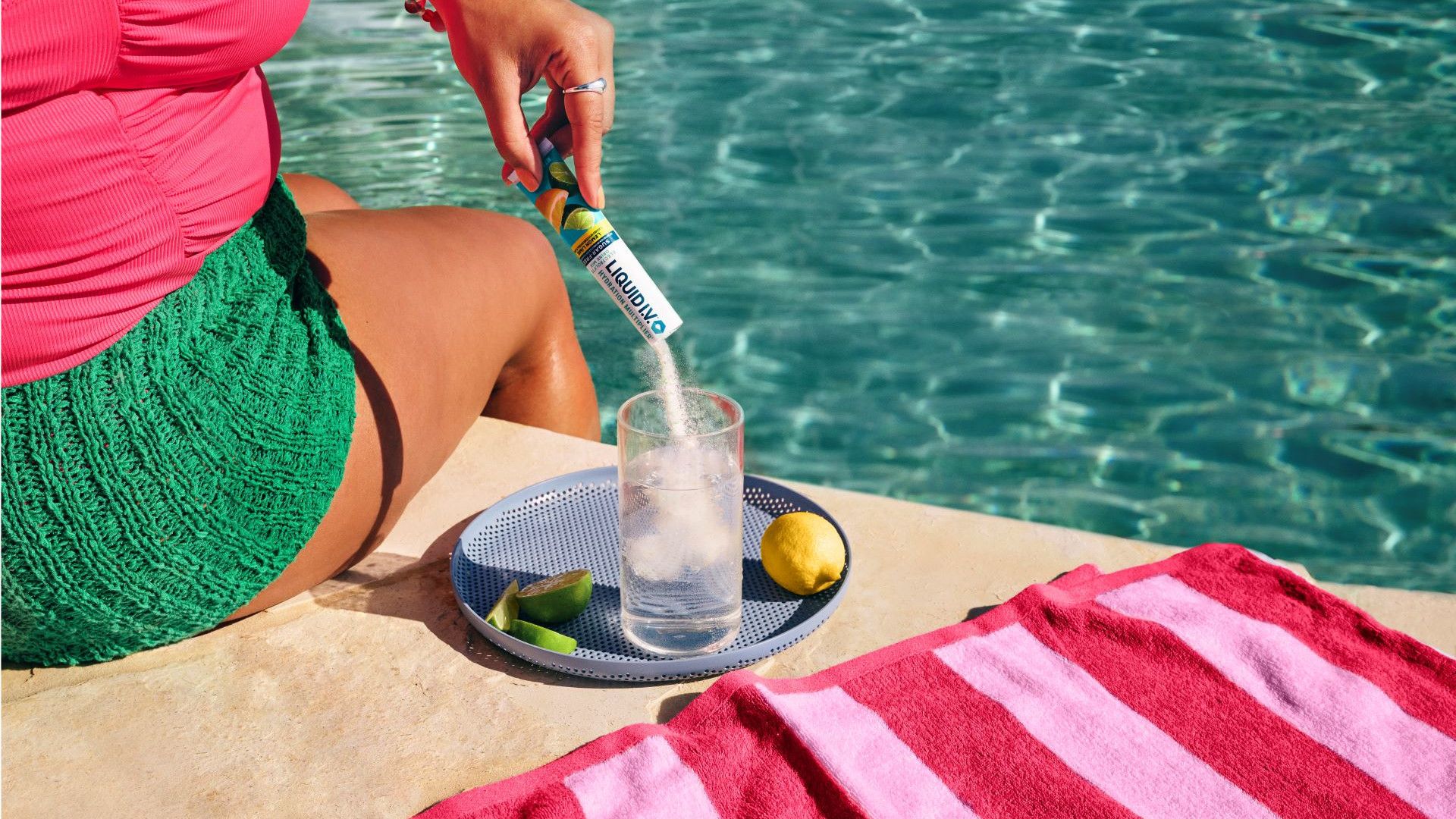 Dame zittend naast een zwembad giet Liquid I.V. hydratatiemix in poedervorm in een glas water.